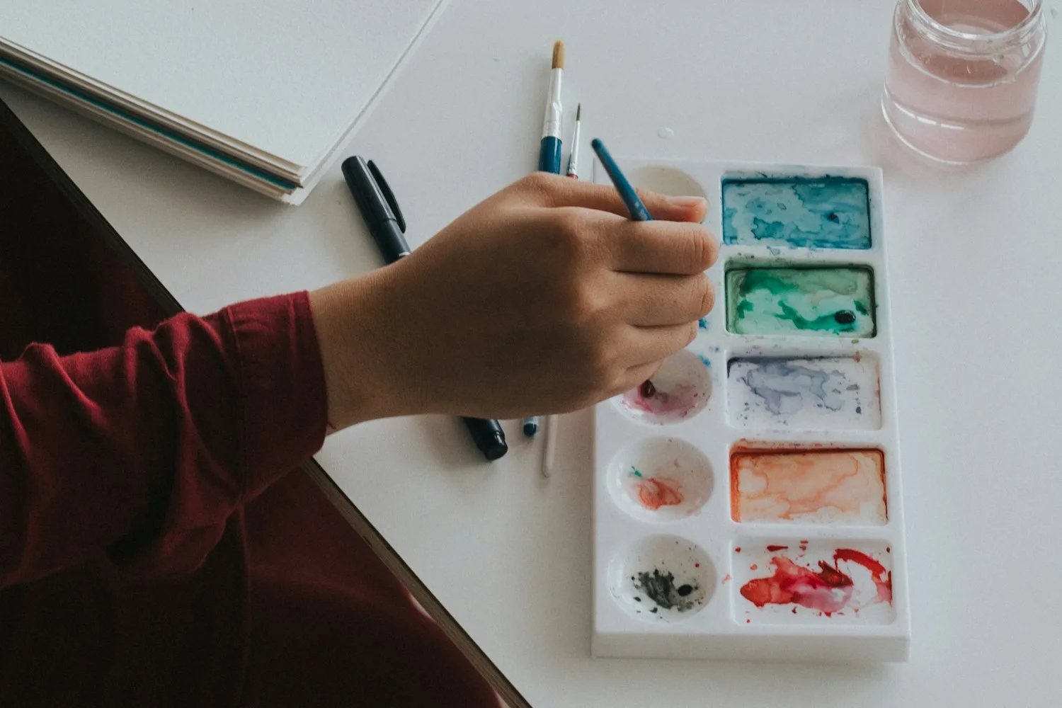 A hand holding a brush above a watercolour palette on a white table