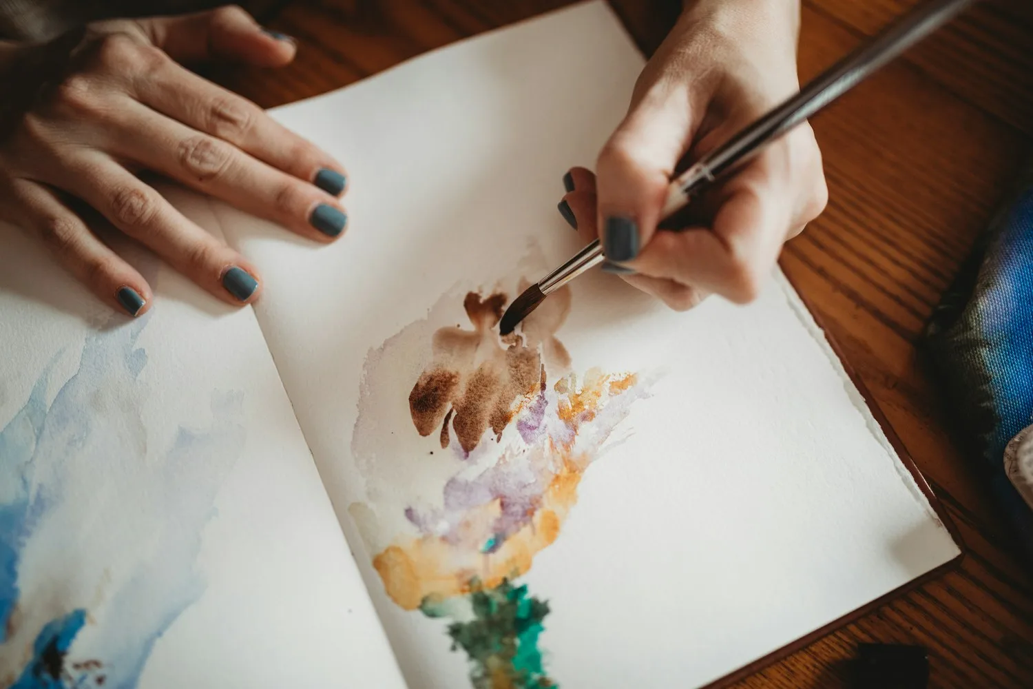 Hands painting loose watercolour shapes in an open journal on a wooden table
