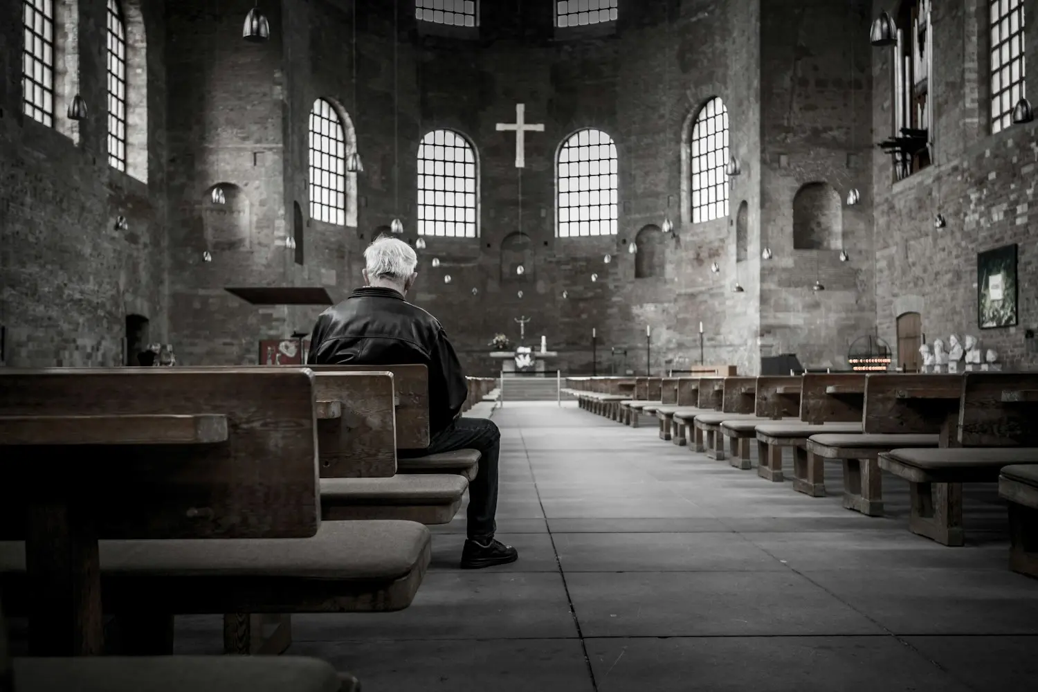 An older man sitting alone on a pew in a quiet brick church, in silent prayer